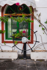 A black ship's wheel with a white mask hangs by a green arched window in Mogan, Gran Canaria. Pink bougainvillea and vines frame white planters on a whitewashed wall. © Aerial Film Studio