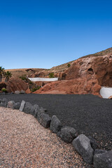 Terraced black lava gravel and basalt stones lead to red tuff cliffs with whitewashed walls in Lanzarote, palm trees rise from a ravine, midday light defines forms. © Aerial Film Studio