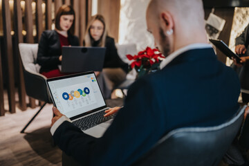 A man in a suit works on a laptop during a business meeting while colleagues discuss in the background. The scene shows a professional workspace with teamwork and a shared screen.