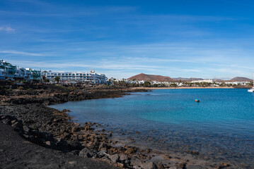 Clear turquoise bay borders dark volcanic rock and resorts in Lanzarote. Rust red cones rise behind the town, palm promenades line the shore, and a small boat floats. © Aerial Film Studio