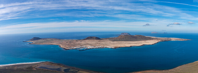 Aerial view from Mirador del Rio shows La Graciosa, Caleta del Sebo, Montana Amarilla, Montana del Mojon, and the Strait of El Rio under wispy clouds in the Canary Islands. © Aerial Film Studio