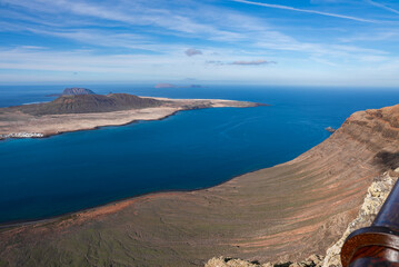 Mirador del Rio in Lanzarote faces La Graciosa and Chinijo Archipelago. Rust cliffs drop to turquoise water. Pale sand flats and volcanic cones sit opposite under high cloud.