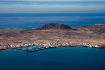 Aerial midday view shows Montana Amarilla above an ochre lava plain. Caleta de Sebo lines a turquoise bay with a marina and breakwater in the Chinijo Archipelago. © Aerial Film Studio