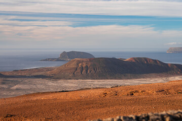 Rust red and black lava ridges lead to a dome caldera, ocean, and Famara cliffs. Late afternoon...
