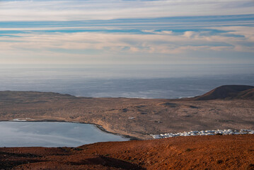 Crescent bay borders ocher volcanic land with whitewashed village near Lanzarote, Canary Islands. Low cinder cones and lava fields meet calm Atlantic under streaked clouds. © Aerial Film Studio