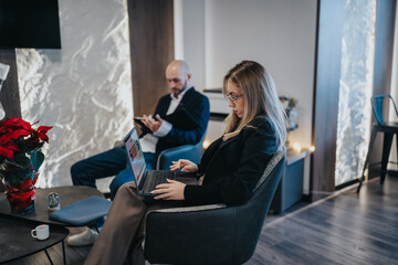 A professional woman uses a laptop in a modern office lounge. In the background, a man works on a tablet, creating a collaborative scene.