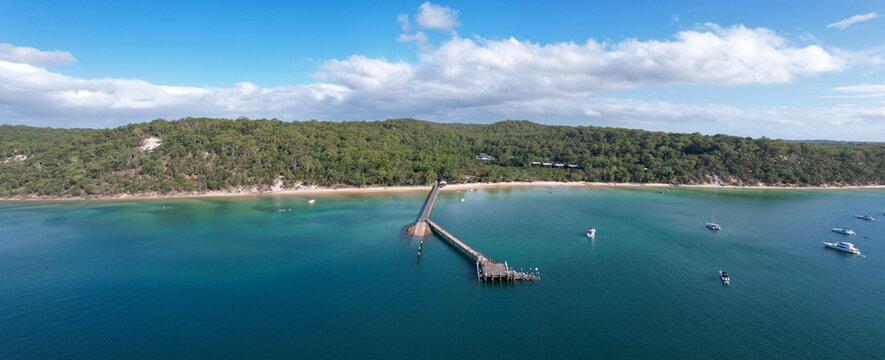 Aerial view of a long pier stretches into the turquoise sea, meeting the sandy shore bordered by lush green forest, Eurong, Queensland, Australia.