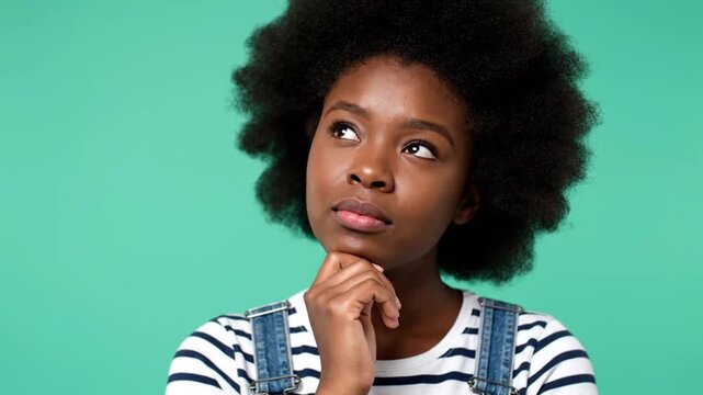 Thoughtful young woman with natural afro hairstyle wearing striped shirt and denim overalls, resting chin on hand and gazing upward with curious expression against teal background.