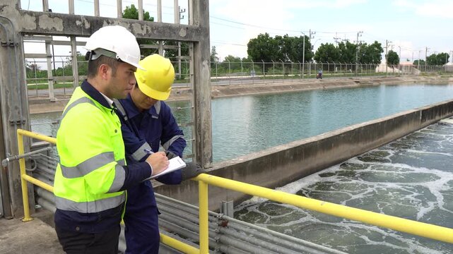 Two Asian engineers discussing maintenance at wastewater treatment plant. They check water quality and pipeline system outdoors. Industrial ecology and environmental management concept.