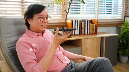 Happy senior businessman talks on smartphone at modern living room. Elderly man connects with AI voice assistant technology at co working space office workplace