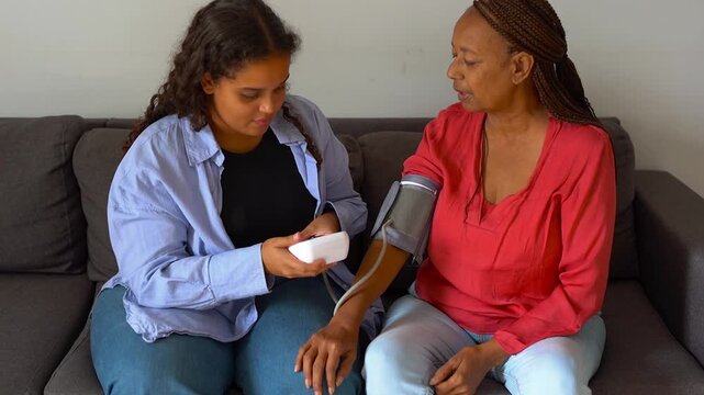 African daughter measuring blood pressure to her mother while looking on monitor - Healthcare, family and lifestyle concept