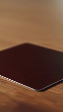 Vertical video: Entering frame burgundy coaster tilting and settling on wood table, casting shadow