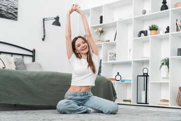 Happy woman stretching arms in bedroom at home, sits cross-legged on floor, resting in a bright,...