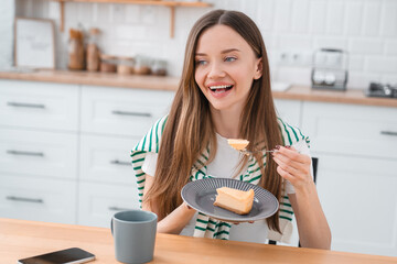 Woman eating a piece of cheesecake, has snack with dessert, savoring the sweet treat. Lifestyle photo, female enjoying birthday cake with cup hot beverage in kitchen home interior.