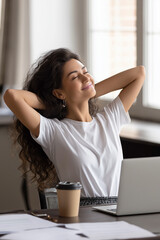 Beautiful Woman relaxing in an office chair with her hands behind her head, laptop and coffee cup on the desk in a bright office setting