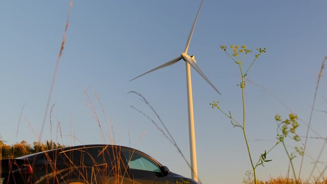 Wind turbine stands above meadow while black car edges frame. Turbine rotates slowly and rotor sweeps arc keeping constant rhythm without pause