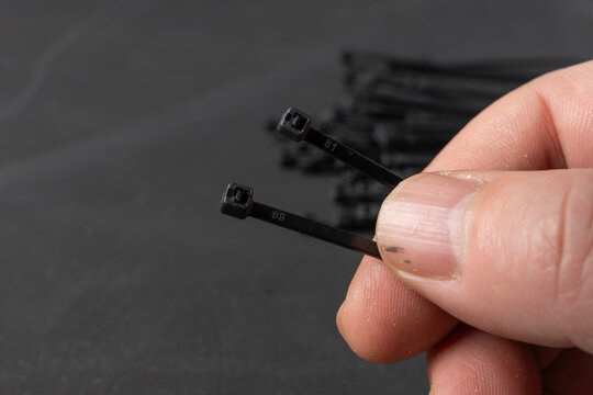Hand holding two black cable ties in front of a blurred pile. Close-up image representing selection, fastening, cable management, and technical work.