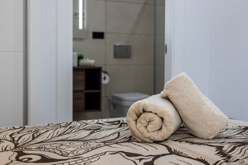 Rolled beige towels on patterned bedspread with view into modern tiled bathroom interior