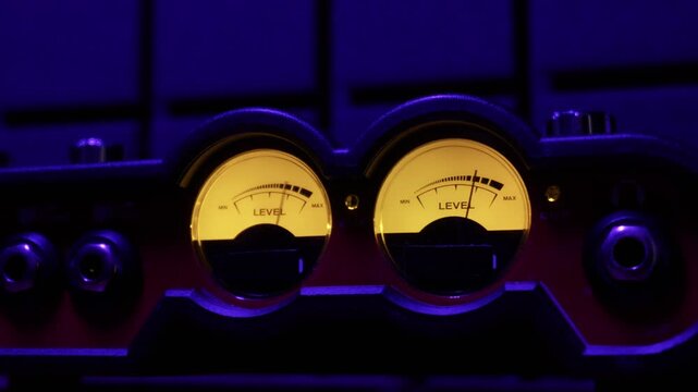 Two analog VU meters on audio equipment measure sound levels in a dark, blue-lit recording studio. Concept of music production, audio engineering, broadcasting, and precise measurement.