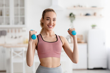 Happy athlete works out at home, exercises for hands and keeps fit during COVID-19 lockdown. Smiling young attractive pretty blonde woman in sportswear lifts dumbbells in minimalist kitchen interior