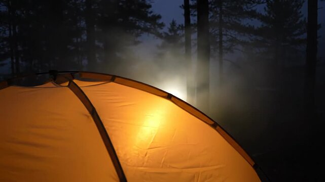 Illuminated tent glows warmly against a dark, misty forest backdrop at night.