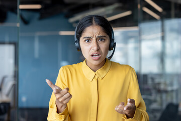 Portrait of a young worried Indian woman sitting in the office wearing a headset and looking at the...