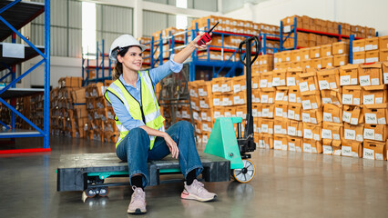Professional female warehouse worker using radio communication and sitting on pallet jack in a storage facility. Team of logistics experts managing inventory and industrial supply chain.