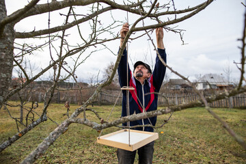 A man hangs a bird feeder on a tree. Man in the garden. A man works in the garden. Garden care. Autumn garden work. Rural housework