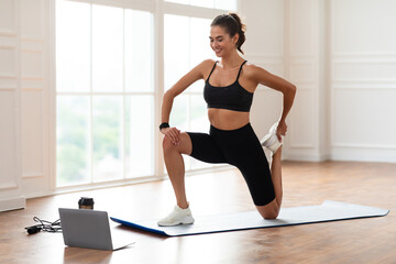 Online Training Session. Happy fit lady in sportswear stretching leg muscles and quads watching online tutorial on pc, exercising in minimal interior living room or fitness studio, looking at screen