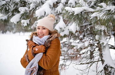 Fototapeta premium Smiling middle aged woman in warm brown sheepskin coat and white hat in winter forest. Beautiful female enjoys snowy nature. Christmas, travel, weekend, lifestyle concept