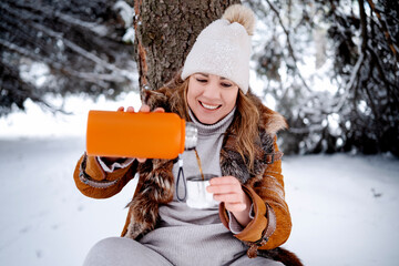 Fototapeta premium Smiling woman pours hot tea from orange thermos in winter forest. Middle aged female sits near tree on snow. Picnic, relax, nature, weekend, wellness concept