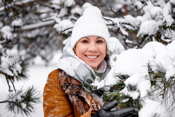 Fototapeta premium Close up portrait of beautiful smiling woman near snowy pine branches. Winter nature beauty. Female in brown coat and white hat. Happiness, relax, lifestyle concept