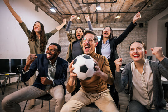 Happy office team watching a football game during a work break, cheering and celebrating together, with one person holding a football, symbolizing unity and work life balance.