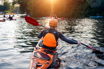 Guy paddles confidently in a bright orange kayak, training on a tranquil river surrounded by lush greenery. Other kayakers are visible nearby under the warm glow of the setting sun