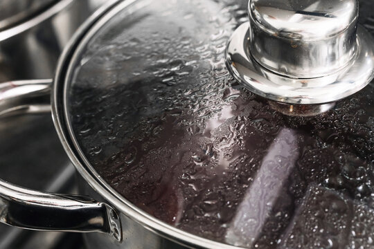 Pot with glass lid covered in steam during boiling process. Cooking soup or broth on stove. Condensation droplets on cookware surface. Home kitchen food preparation scene closeup.