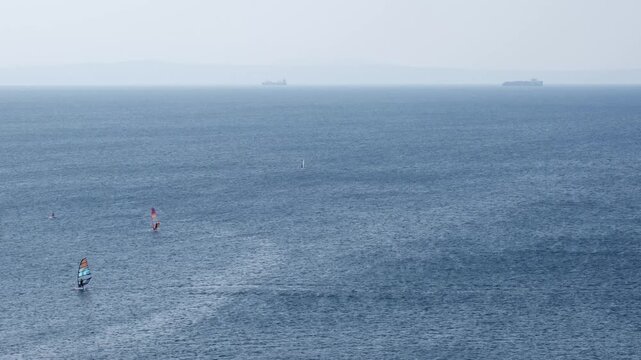 Windsurfers ride waves on the open Adriatic Sea. High-angle seascape with large anchored container or cargo ships in the background. Windy summer day in the Kvarner Bay, Croatia.