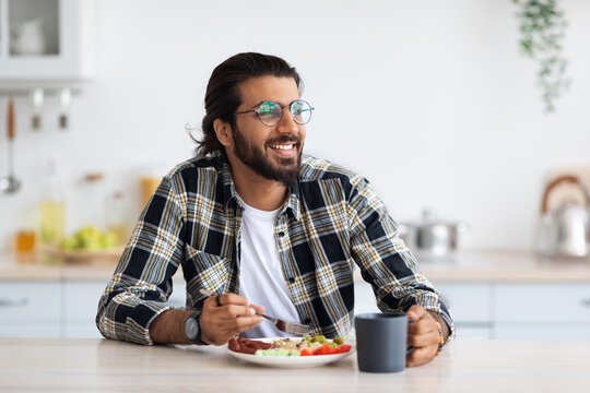 Happy middle-eastern young bearded man in casual outfit and stylish glasses enjoying healthy lunch at home, sitting at kitchen table, eating breakfast, looking at copy space, healthy deit concept