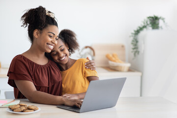 Cute black mother and kid using laptop together, sitting at kitchen table with homemade pastry,...