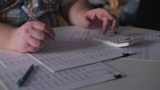 Man Calculates Expenses On Desk, Man Reviews Bills And Calculates Expenses During Evening Hours, Caucasian Man Uses Calculator And Pen To Tally Bills In Low Evening Light