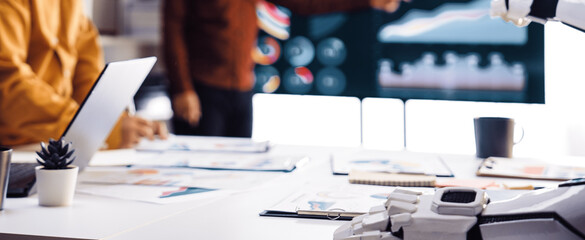 A dynamic business meeting showcases the integration of advanced technology with a robot hand assisting team members. Innovative visuals enhance strategic discussions.Noogenesis