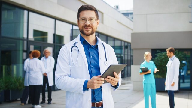 Confident Caucasian male doctor standing outside hospital. Medical specialist holding tablet. Background colleagues discussing cases. Outdoor clinic area hosting professional team.