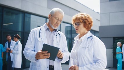 Two senior doctors standing outside hospital building reviewing tablet and holding focused posture. Caucasian specialists analyzing patient information together. Medical staff in background.