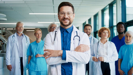 Confident Caucasian male doctor standing in bright corridor crossing arms. Smiling medical leader inspiring diverse team behind him. Showing assurance during busy hospital day. People feeling proud.