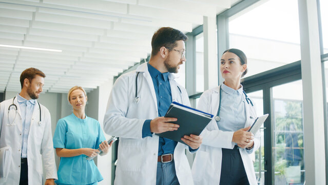 Medical staff walking through bright clinic hallway talking with each other. Focused male doctor reviewing notes while colleague discuss plan. Nurse joining conversation and sharing thoughts.