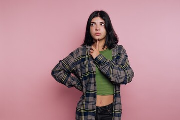 Young woman in plaid shirt pondering against pink background.