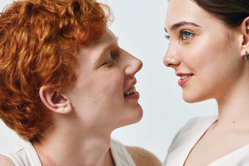 Fototapeta premium Portrait of young couple with joyful expressions looking at each other closeup. Smiling man with red hair and woman with blue eyes in intimate moment on light background.