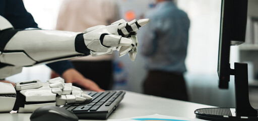 A close-up shot of a robotic arm interacting with a computer keyboard, showcasing modern technology in a business environment, with professionals collaborating in the background.Noogenesis