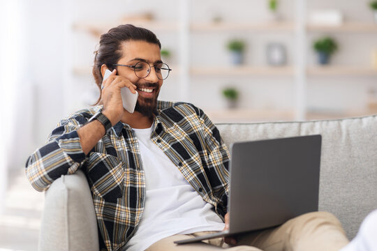 Positive long-haired, bearded arab guy freelancer sitting on sofa with lapto, having phone coversation, typing on computer keyboard, working from home, looking at screen and smiling, copy space