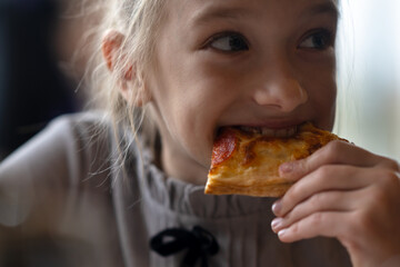 Naklejka premium A close-up portrait of a girl biting into a pizza while holding it in her hand. A 9-year-old child with blonde hair eats pizza. High quality photo
