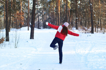A girl in a bright jacket, scarf and hat walks through the winter forest. Rear view. Walking through the winter forest. Snowy cold weather. Active recreation. Woman in a red jacket in the forest
